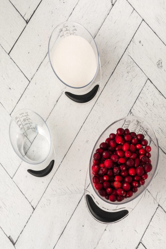 Overhead view of water, sugar, and fresh cranberries measured out in clear cups on a white wood surface.