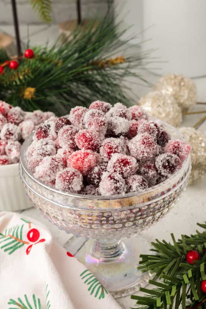 Finished sugared cranberries in a glass bowl, sparkling and festive with Christmas greenery in the background.