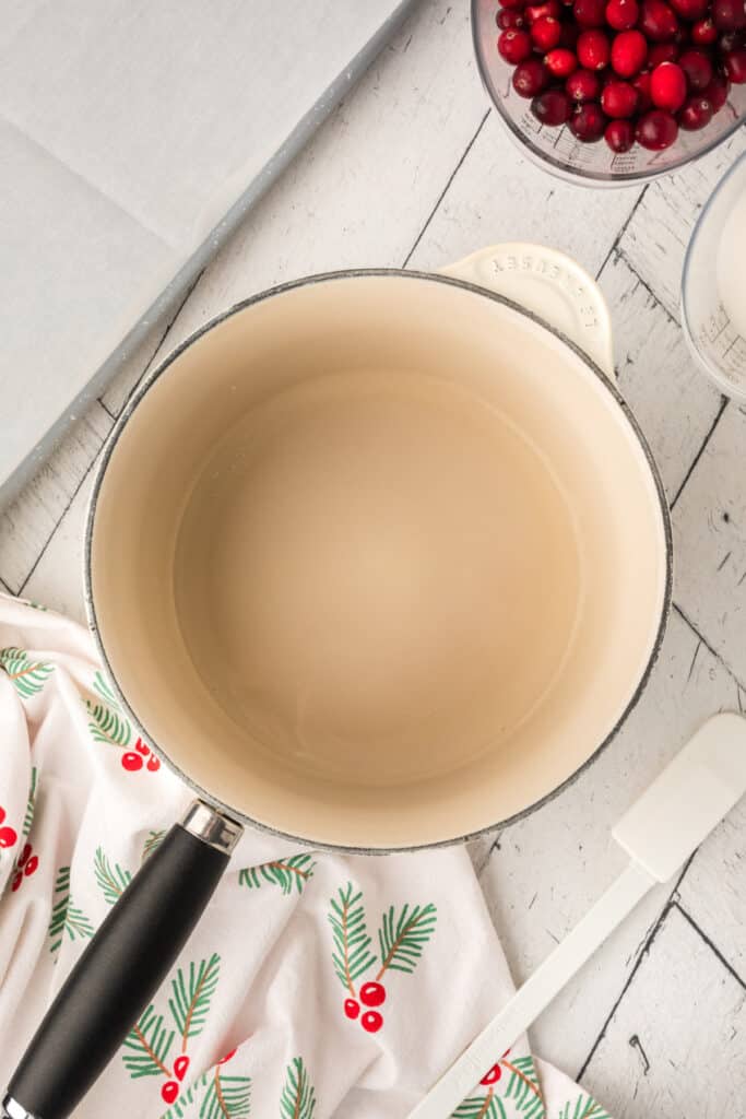 Saucepan filled with water and sugar mixture on a festive towel, cranberries and utensils nearby.