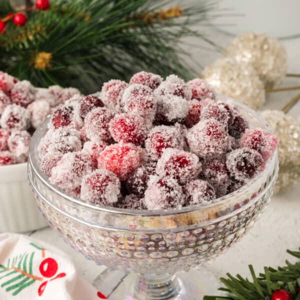 Square crop of sugared cranberries in a glass bowl with pine greenery and holiday decorations in the background.