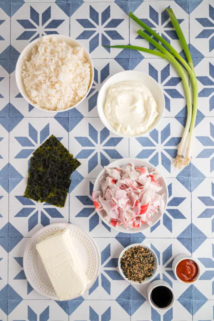 Overhead shot of sushi bake ingredients on a blue and white tiled surface, including sushi rice, crab sticks, cream cheese, mayonnaise, soy sauce, sriracha, furikake, and green onions.