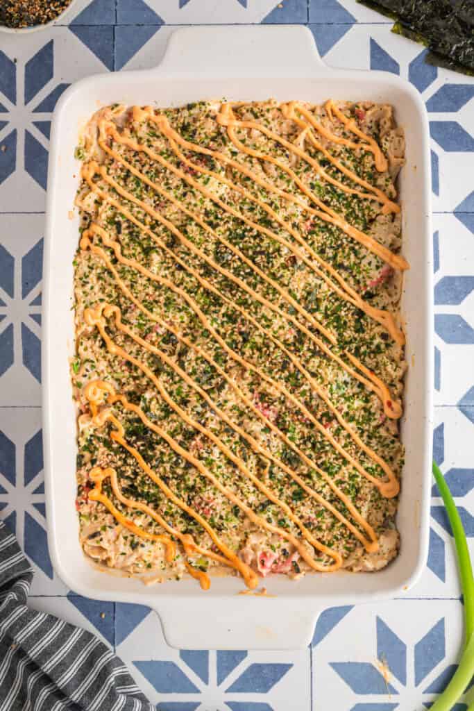 Finished sushi bake in a white baking dish topped with furikake and drizzled with sriracha mayo in neat diagonal lines.