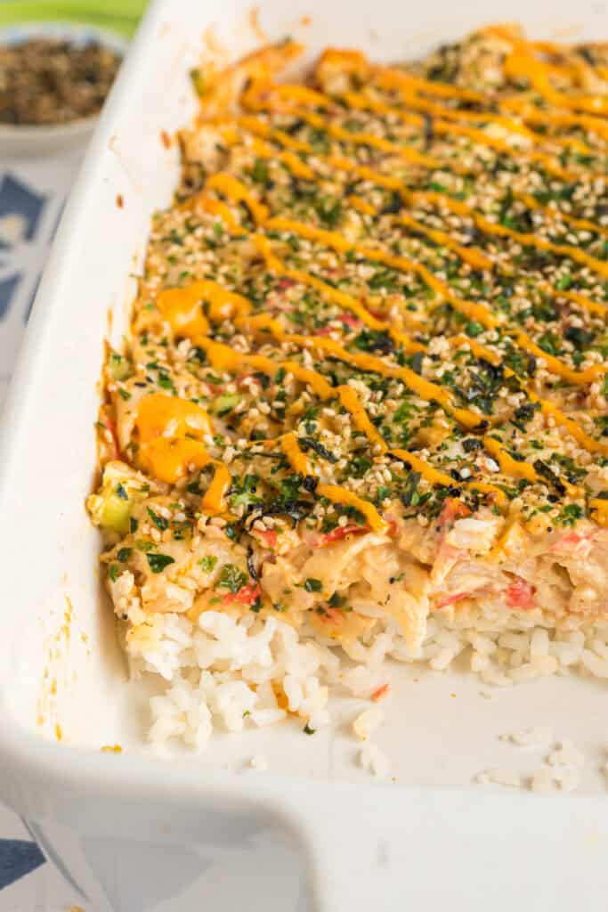 Close-up of a partially scooped sushi bake showing layers of rice, creamy crab filling, and spicy mayo topping.
