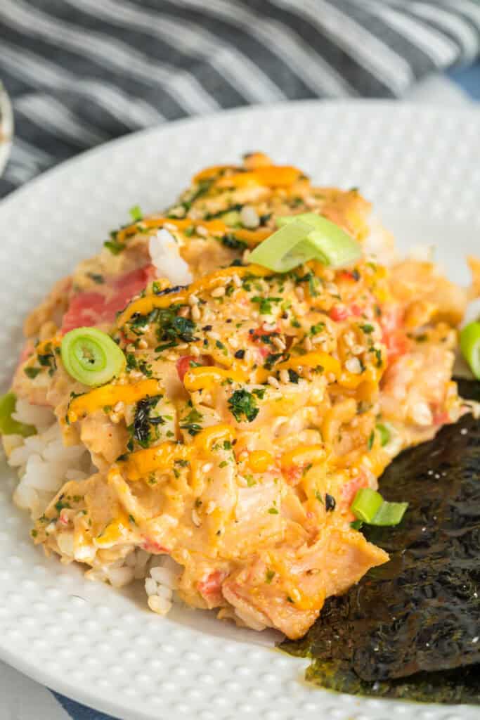 Plated serving of sushi bake on a white dish with green onions, seaweed, and furikake garnish.