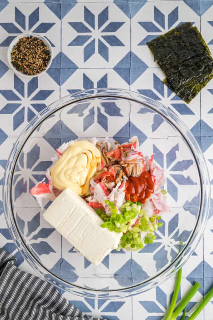 Ingredients for sushi bake in a clear mixing bowl before stirring, with imitation crab, cream cheese, kewpie mayo, sriracha, soy sauce, and green onions.