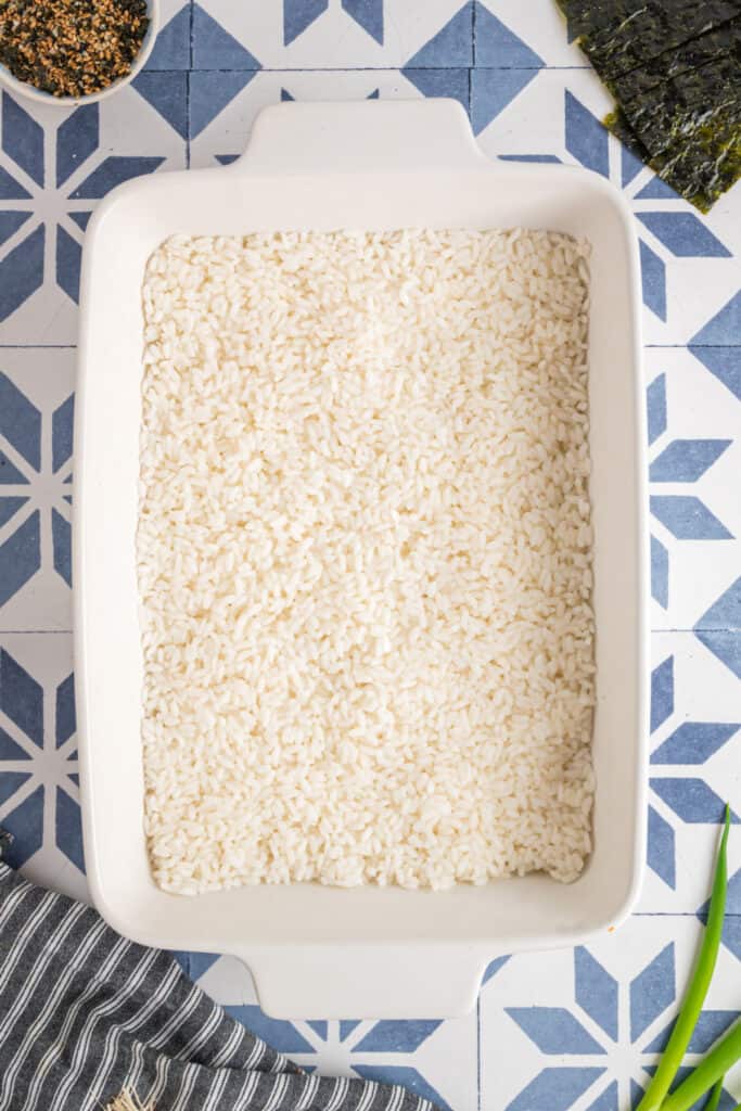 Baking dish with a layer of cooked sushi rice spread evenly across the bottom, ready for topping.
