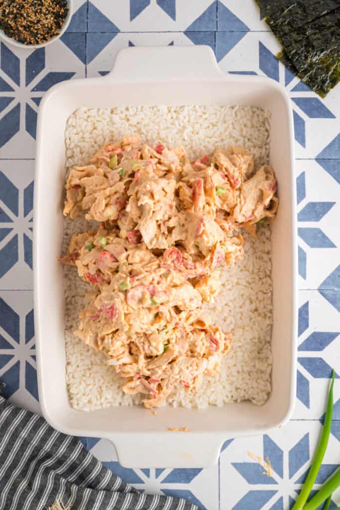 Sushi bake being assembled with the creamy crab mixture spooned over the rice layer in a baking dish.