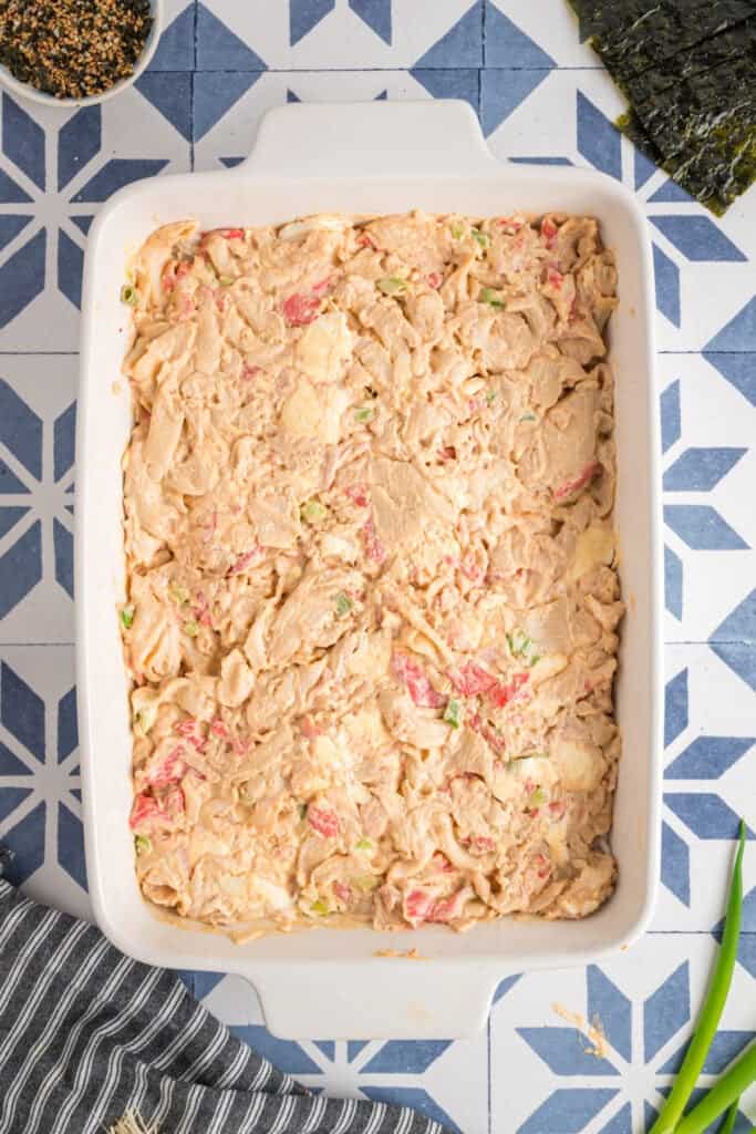 Sushi bake filling evenly spread over the rice in a casserole dish, showing creamy texture and bits of crab and green onion.