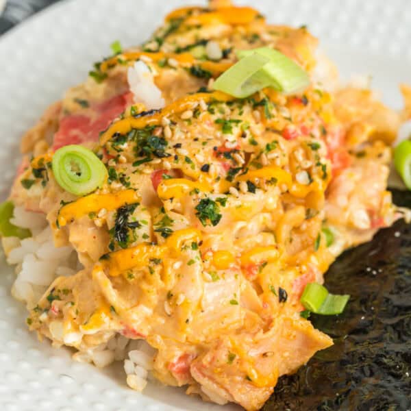 Close-up square image of sushi bake on a plate with creamy crab filling, furikake, and spicy mayo drizzle.