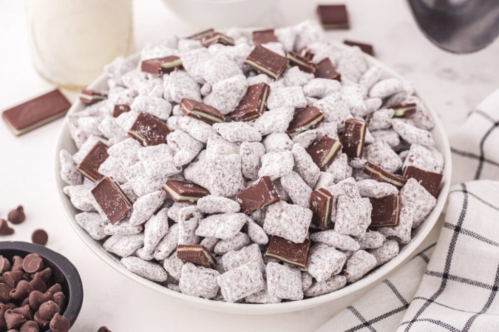 Bowl of Thin Mint Muddy Buddies topped with chopped Andes mints, with chocolate chips and a glass bottle of milk in the background.