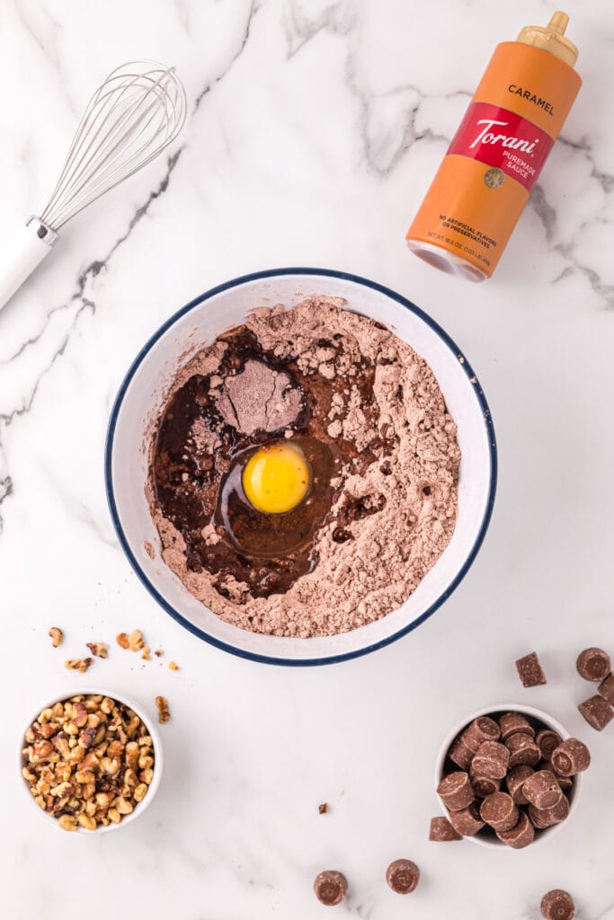 Mixing bowl with brownie mix, egg, water, and oil before being stirred, surrounded by walnuts, Rolos, caramel sauce, and a whisk on a marble surface.