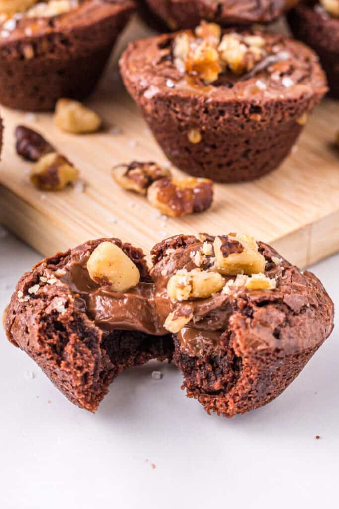 Close up of a turtle brownie bite broken open to show melted chocolate and chopped walnuts in the center, with more brownie bites behind it.