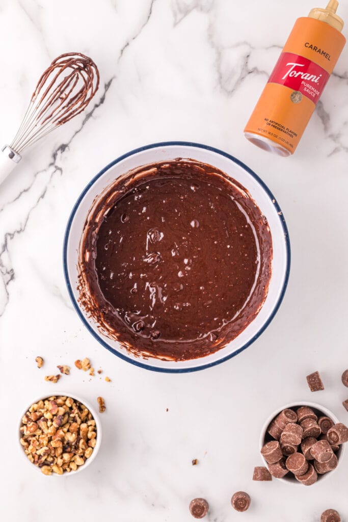 Smooth brownie batter mixed in a white bowl with a whisk and caramel sauce bottle nearby, with walnuts and Rolos scattered around.