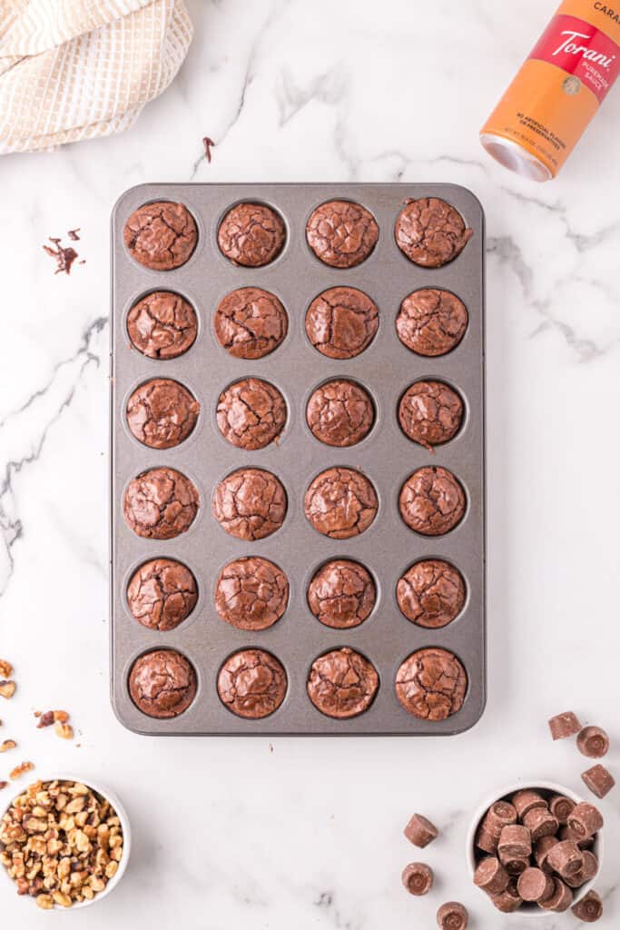 Freshly baked brownie bites still in the mini muffin pan, with cracked tops and ingredients scattered around.