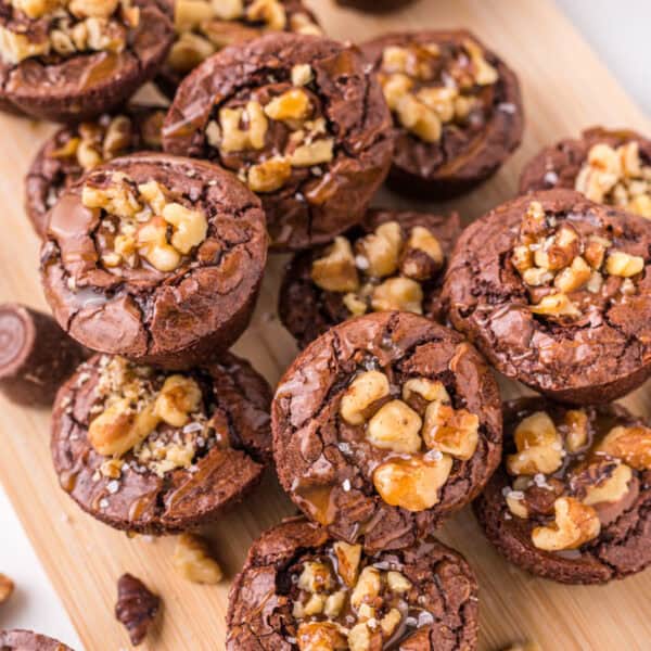 Pile of turtle brownie bites on a wooden board, topped with chopped walnuts and caramel drizzle.