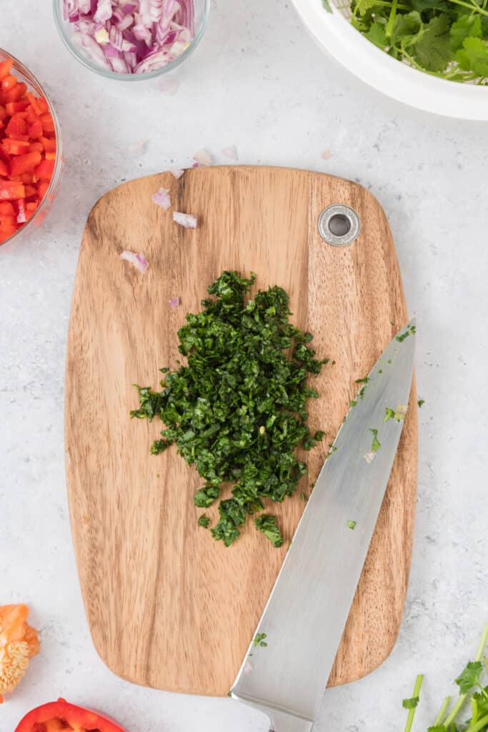 Chopped cilantro on a wooden cutting board with a chef’s knife, diced onions and peppers nearby.