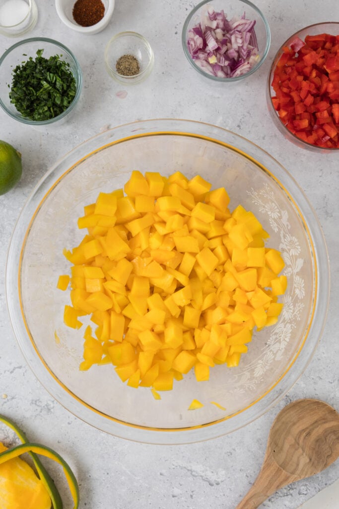Large glass bowl filled with diced mango, surrounded by small bowls of cilantro, onion, pepper, and seasonings.