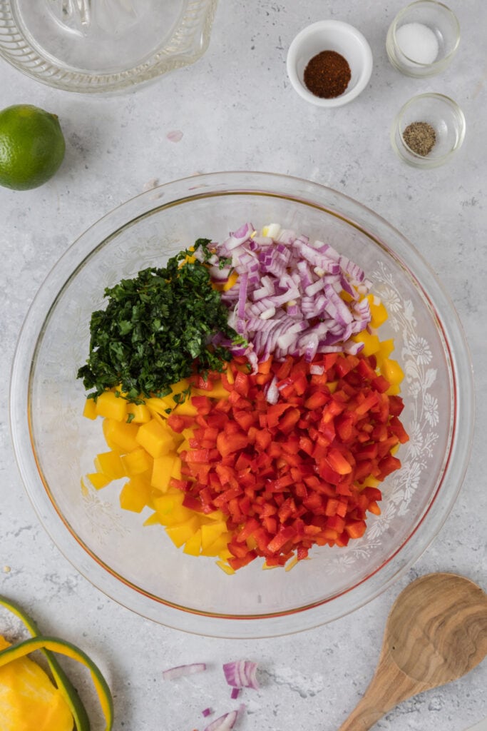 Glass bowl with diced mango topped with diced red pepper, red onion, and cilantro before mixing.