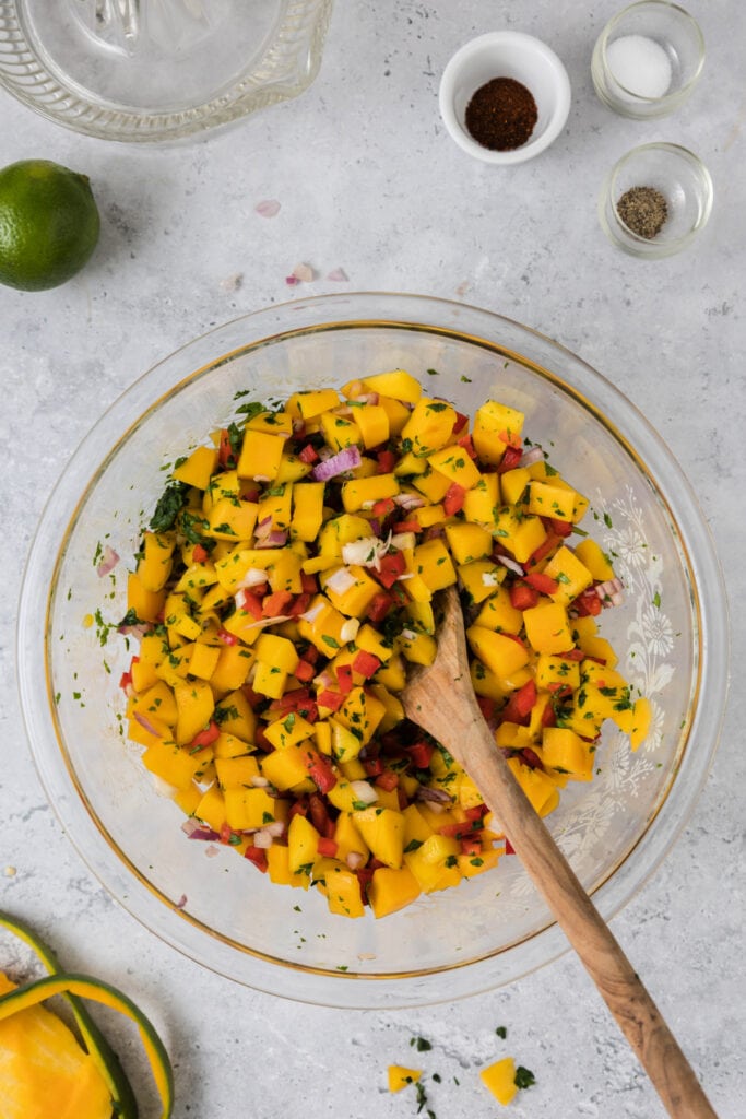 Mango salsa mixture in a large glass bowl being stirred with a wooden spoon.