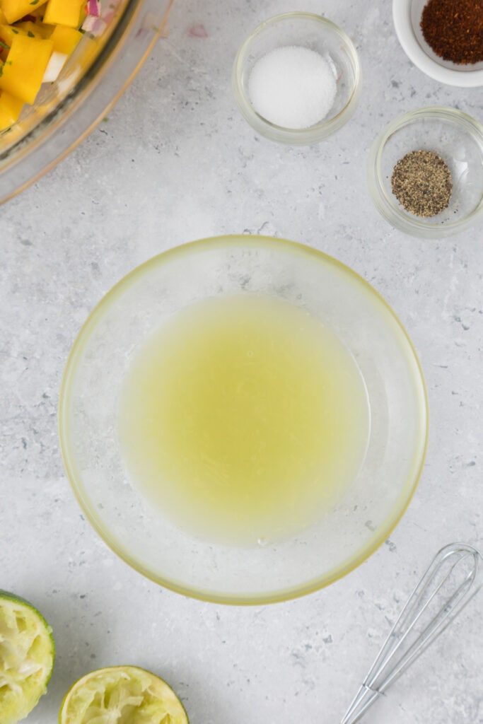 Small bowl filled with fresh lime juice surrounded by lime halves, whisk, and seasonings.