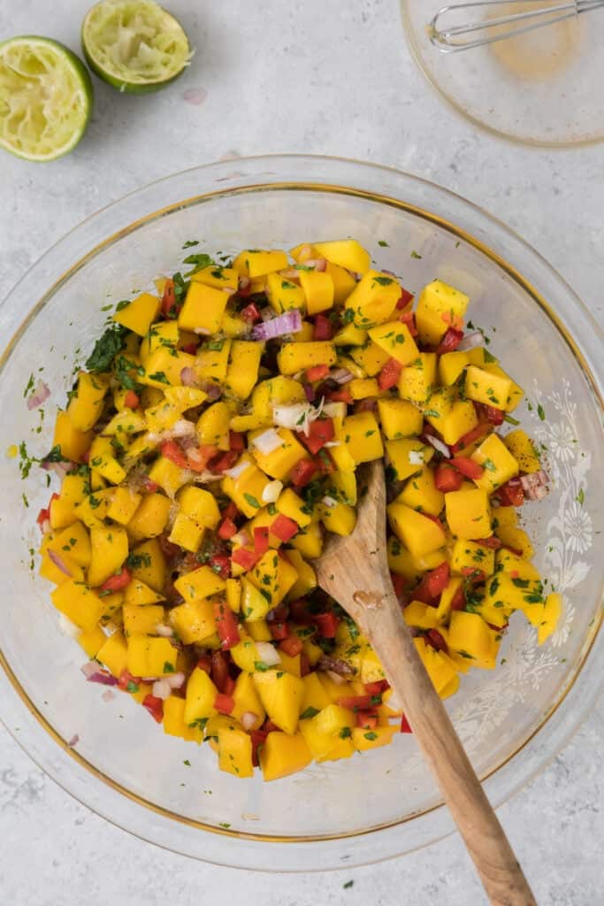 Mixed mango salsa in a large bowl with lime halves and seasoning bowls nearby.