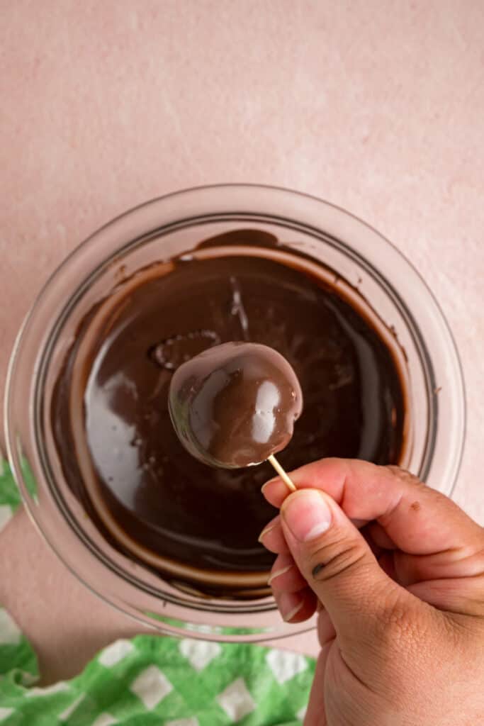 buttercream ball coated in chocolate held on a toothpick above the bowl