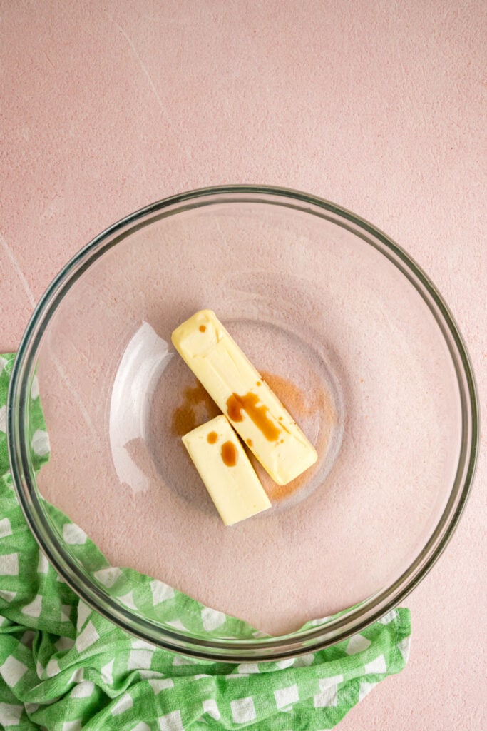 sticks of butter and vanilla extract in a glass mixing bowl on a pink background