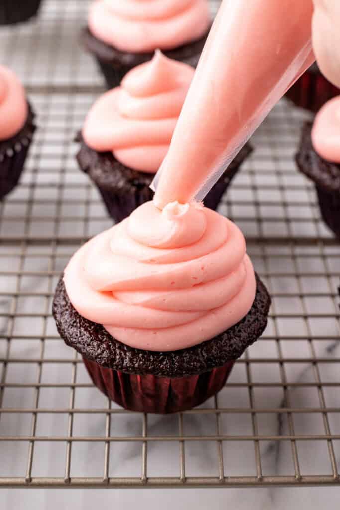 Cherry buttercream frosting being piped onto a chocolate cupcake on a cooling rack
