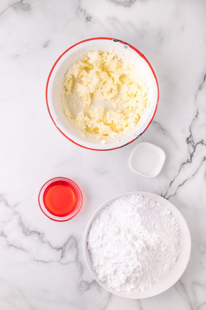 Overhead view of a bowl of whipped butter, a small bowl of red cherry juice, and a bowl of powdered sugar on a marble surface