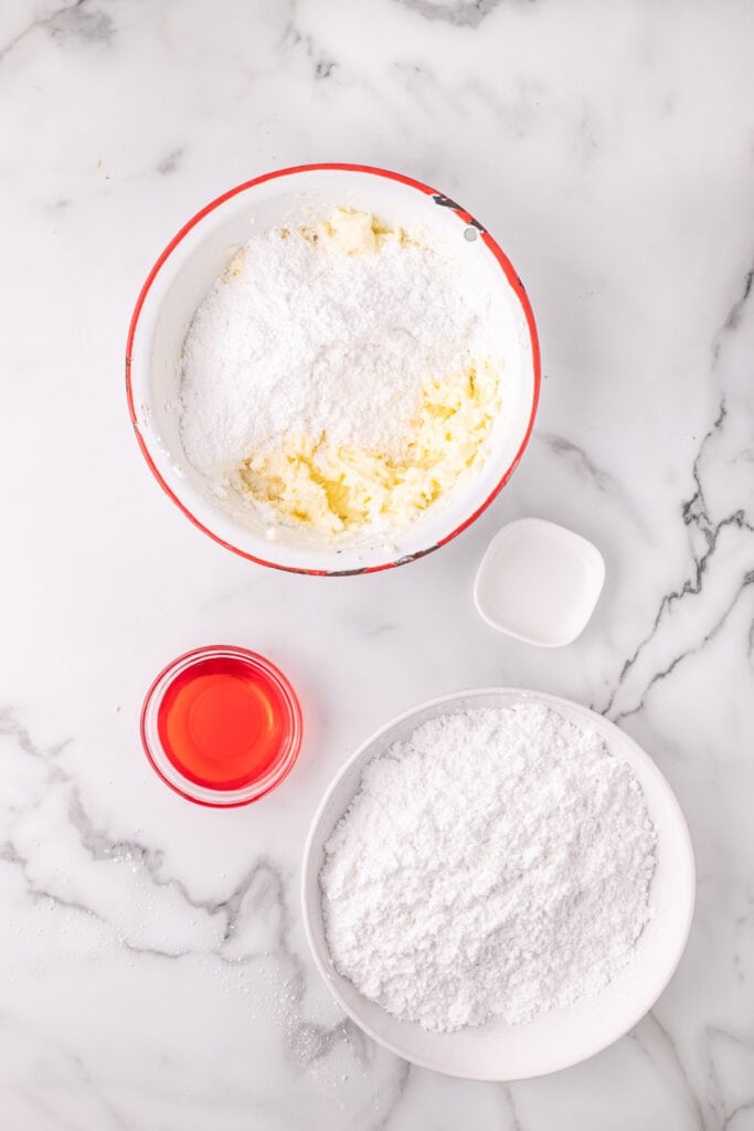 Overhead view of powdered sugar added on top of whipped butter in a mixing bowl with cherry juice nearby