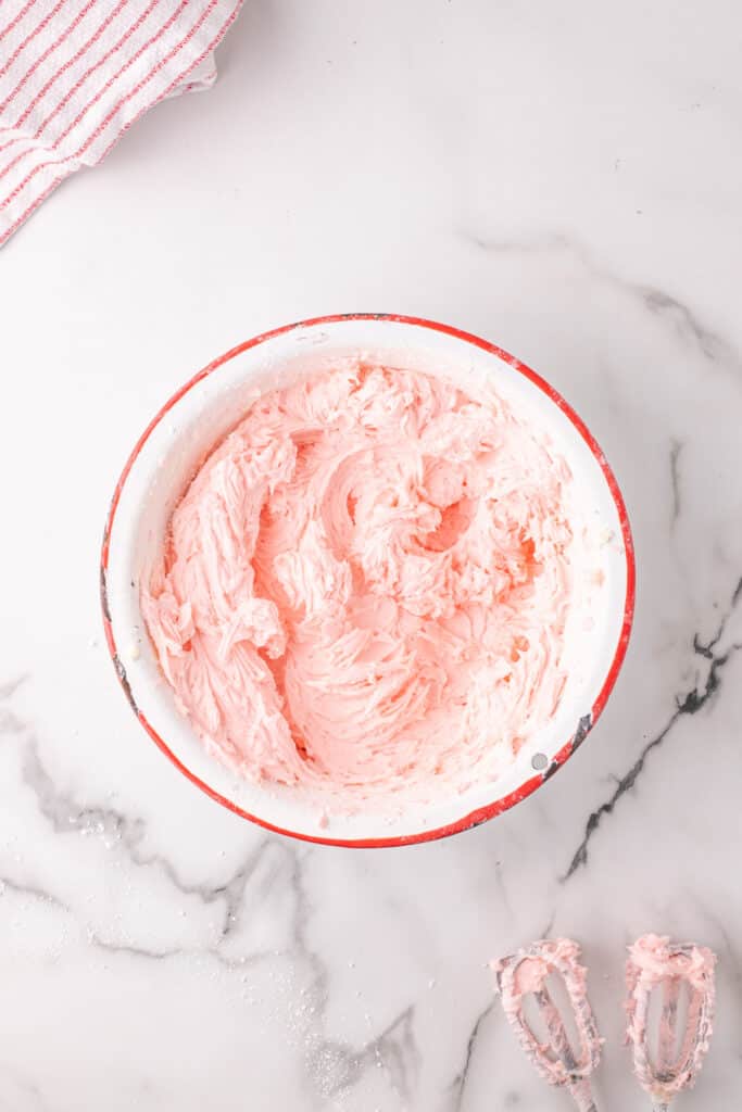 Overhead view of a bowl filled with pale pink cherry buttercream frosting on a marble surface