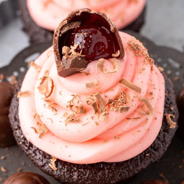 Square image of a chocolate cupcake topped with pink cherry buttercream and a halved chocolate covered cherry