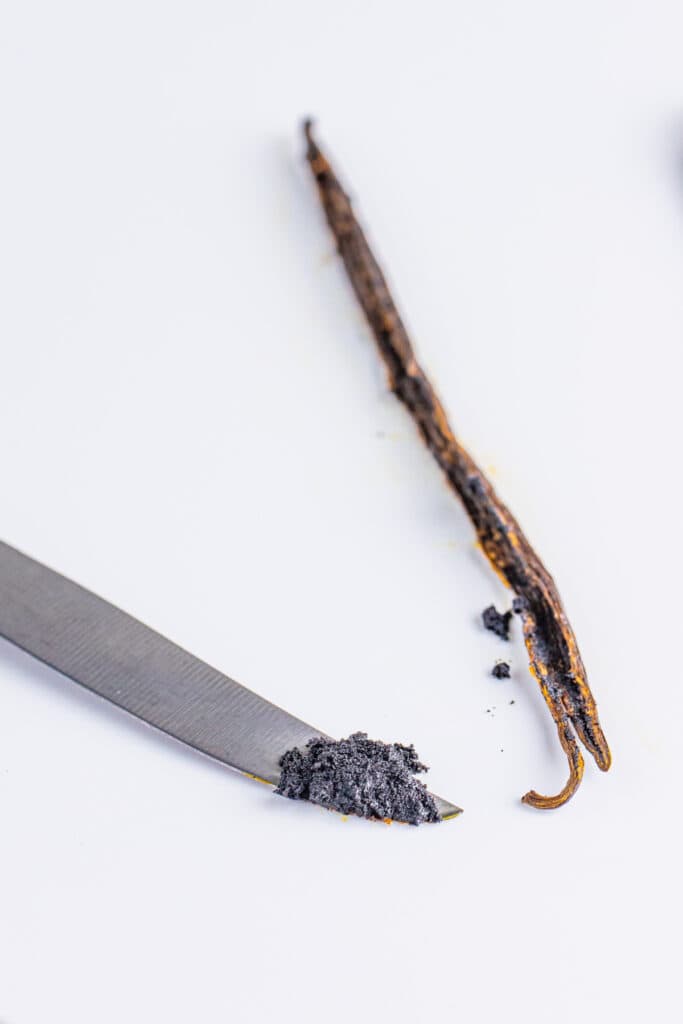 split vanilla bean with scraped vanilla seeds on the tip of a knife on a white surface