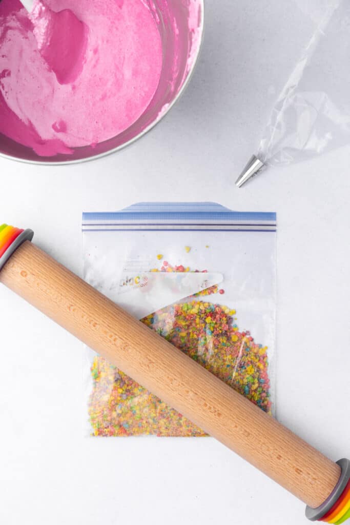 Pink macaron batter in a mixing bowl, piping bag fitted with a round tip, and crushed Fruity Pebbles in a zip top bag with a rolling pin