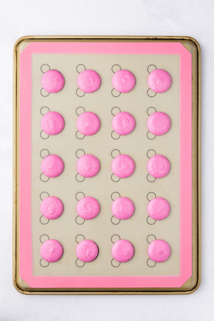 Pink macaron shells piped onto a silicone baking mat in evenly spaced circles before resting