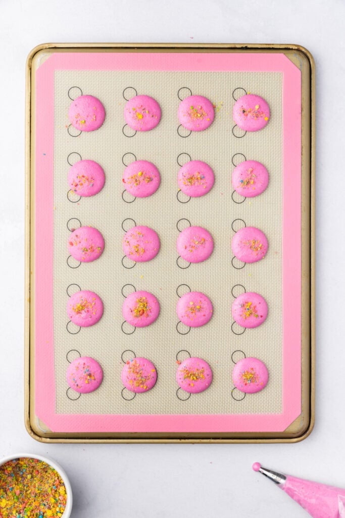 Pink macaron shells on a baking mat topped with crushed Fruity Pebbles before baking