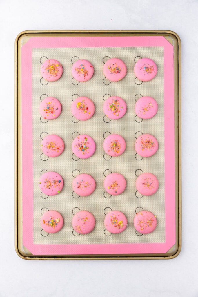 Pink macaron shells baked and set on a silicone mat with cereal topping visible