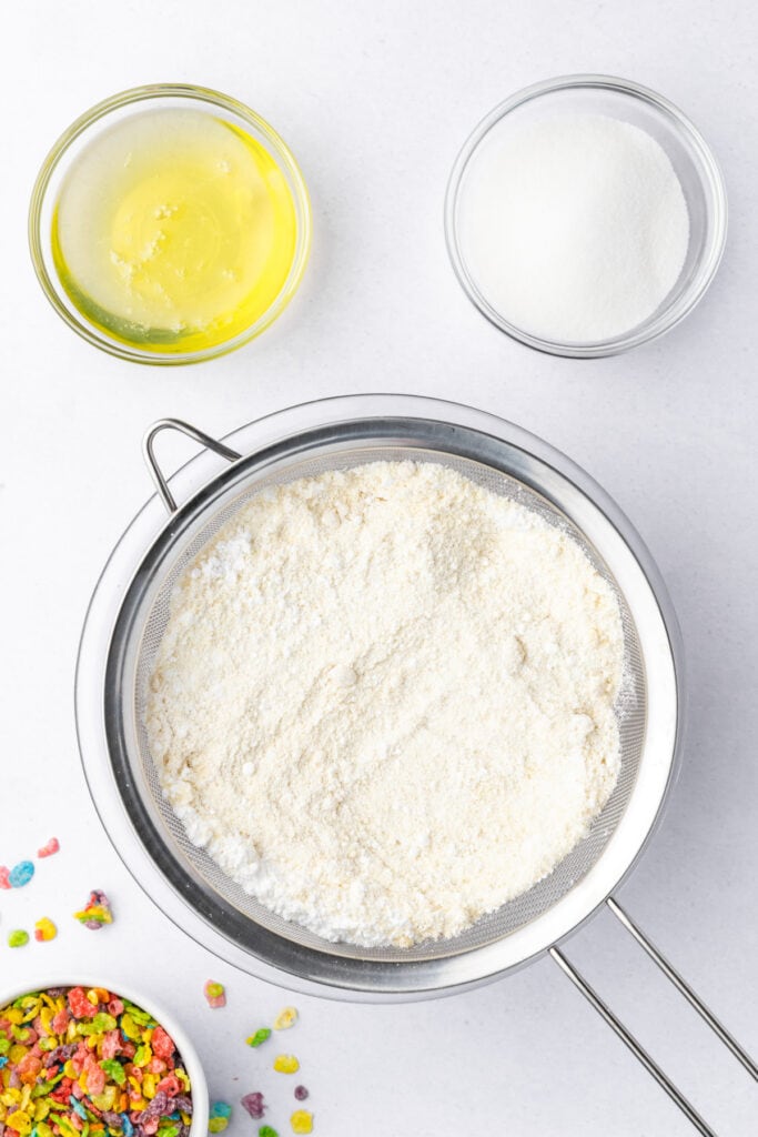 Overhead view of sifted almond flour and powdered sugar in a mesh sieve, with egg whites and granulated sugar measured in small bowls