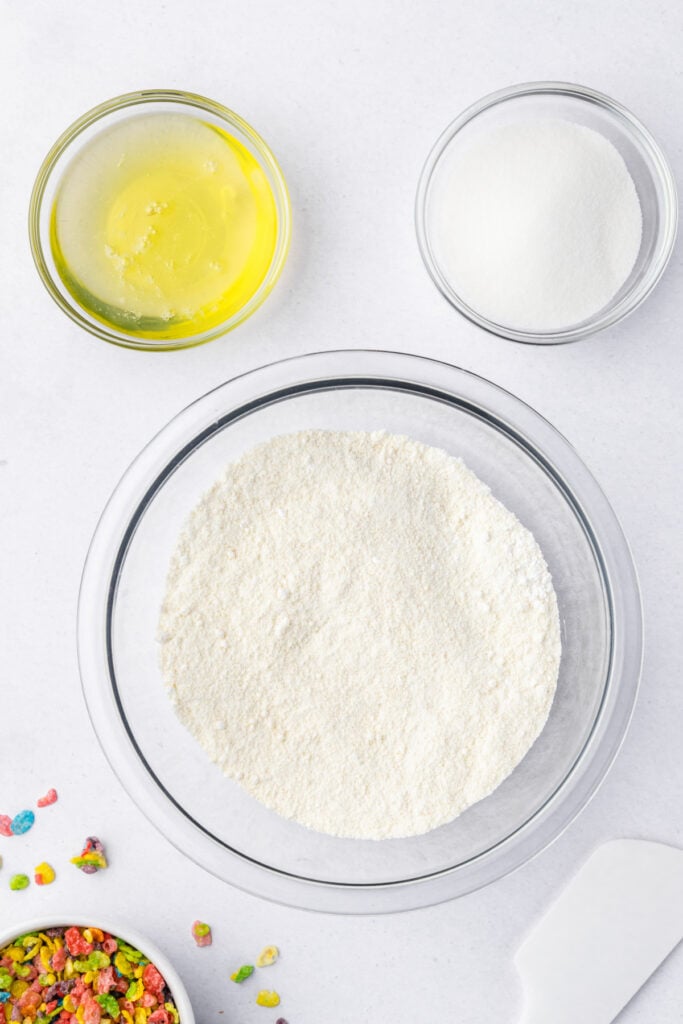 Almond flour and powdered sugar sifted into a clear mixing bowl, with egg whites and sugar measured above