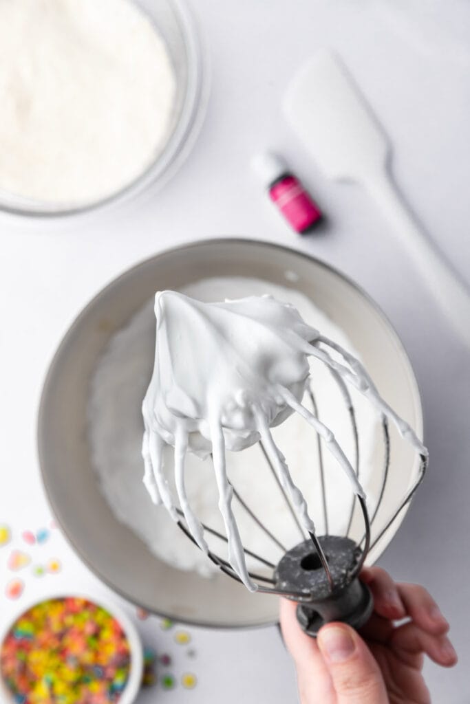 Fully whipped white meringue in a stand mixer bowl with gel food coloring nearby