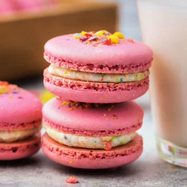 Two stacked pink Fruity Pebbles macarons with speckled filling, crushed cereal on top, and a glass of milk in the background