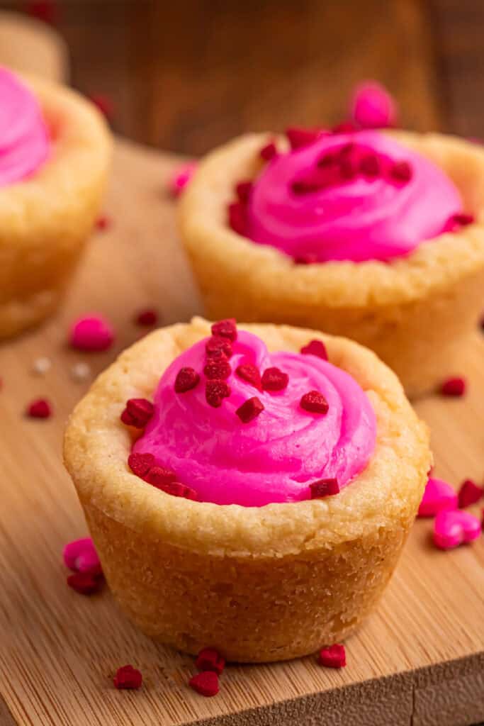 Valentine sugar cookie cup filled with bright pink frosting and topped with red heart sprinkles on a wooden board