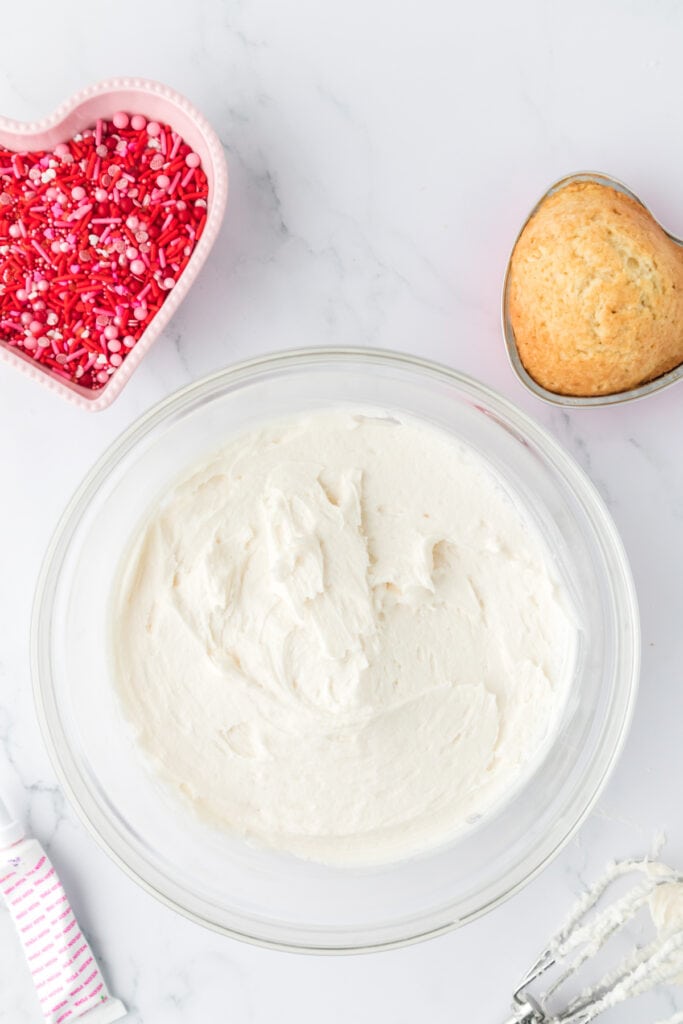 Large glass bowl filled with smooth white frosting, with a heart shaped pan of baked vanilla cake and a bowl of Valentine sprinkles nearby
