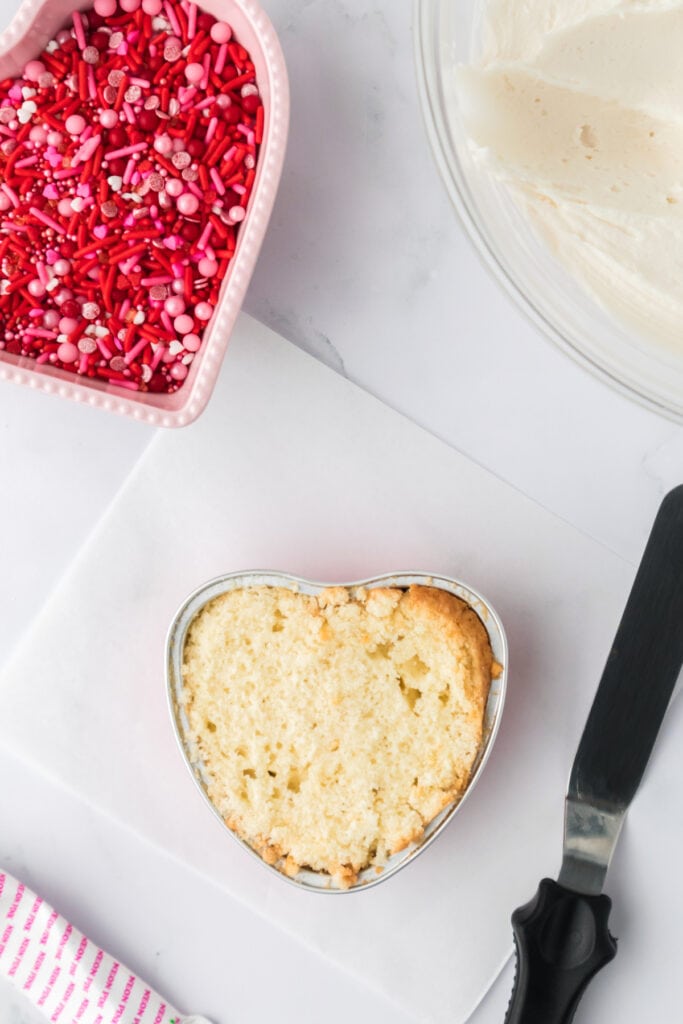 Heart shaped vanilla cake with the top leveled, sitting on a white cutting board next to a spatula
