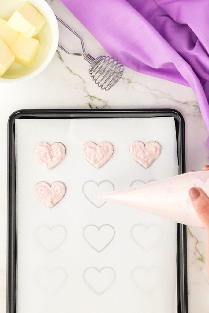 piping heart shaped macaron batter onto parchment paper using a heart template