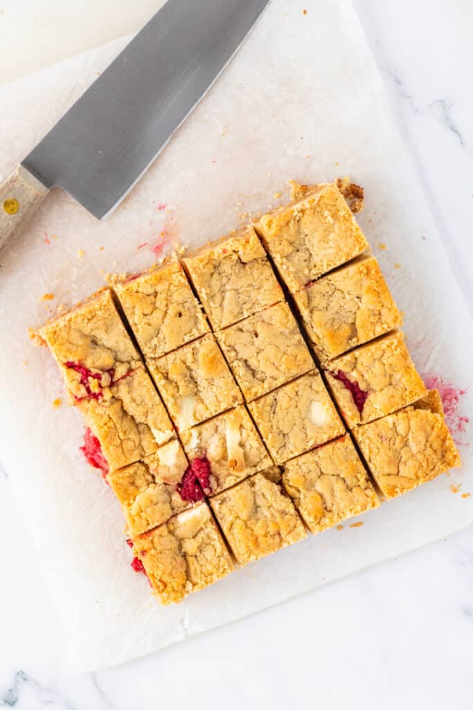 Overhead view of white chocolate raspberry blondies cut into squares on parchment paper with a knife nearby
