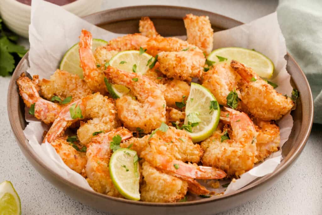 Cooked coconut shrimp served in a bowl lined with parchment paper and garnished with lime wedges and chopped cilantro.