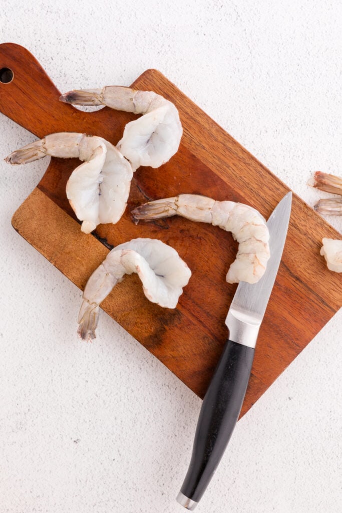 Raw shrimp butterflied on a wooden cutting board with a knife beside them.