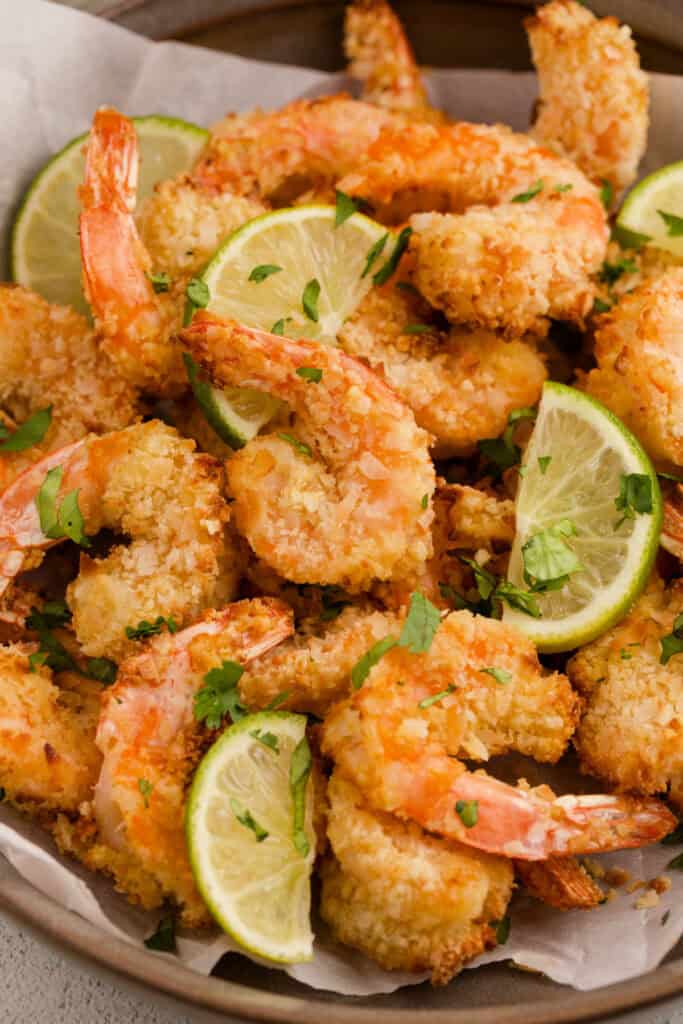 Close up of crispy coconut shrimp with lime wedges and chopped cilantro in a serving bowl.