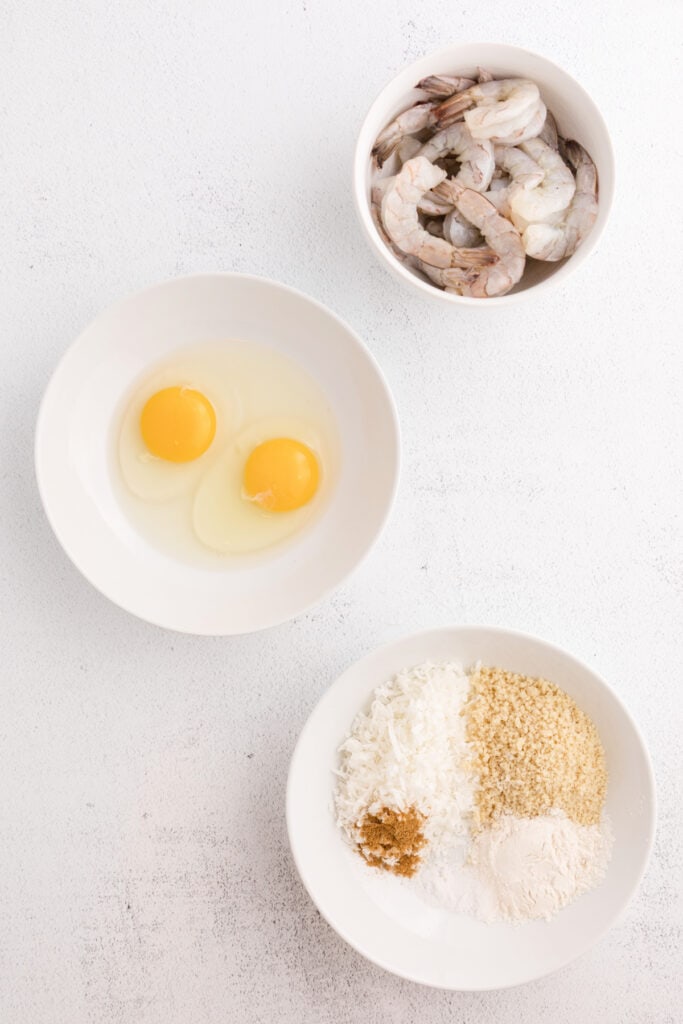 Bowls with raw shrimp, two eggs, and a plate of coconut, panko, flour, and spices on a white surface.